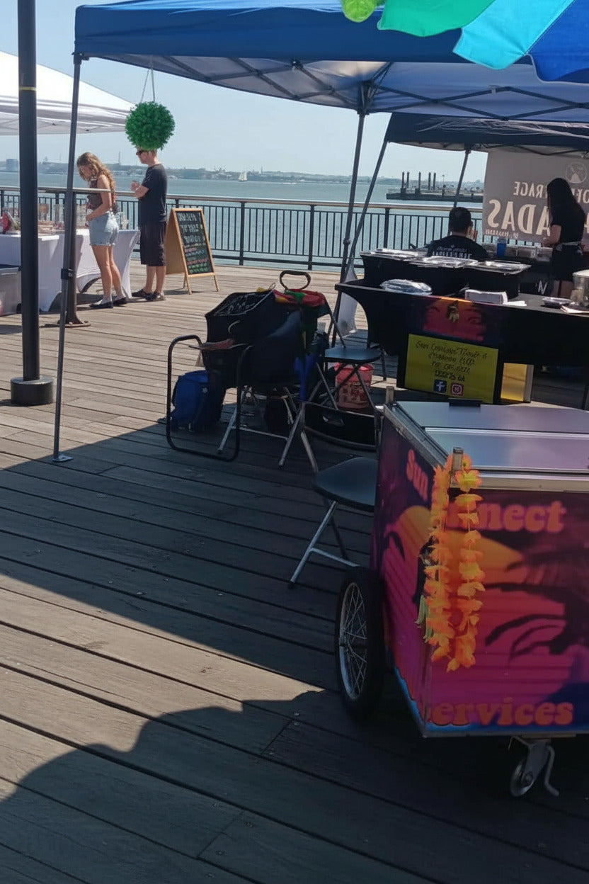 A waterfront event scene with a blue canopy tent, wooden dock flooring, people standing and sitting, and a colorful food cart from Sun Connect Foods & Services in the foreground.