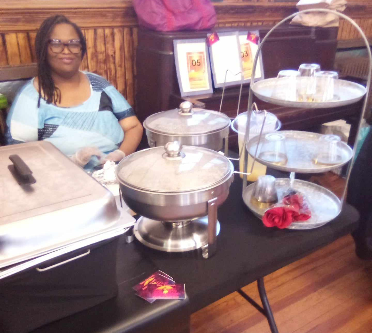 A Sun Connect Foods & Services LLC representative sits behind a buffet-style food table with stainless steel chafing dishes, serving trays, and glass cups in an indoor setting for Business function catering.