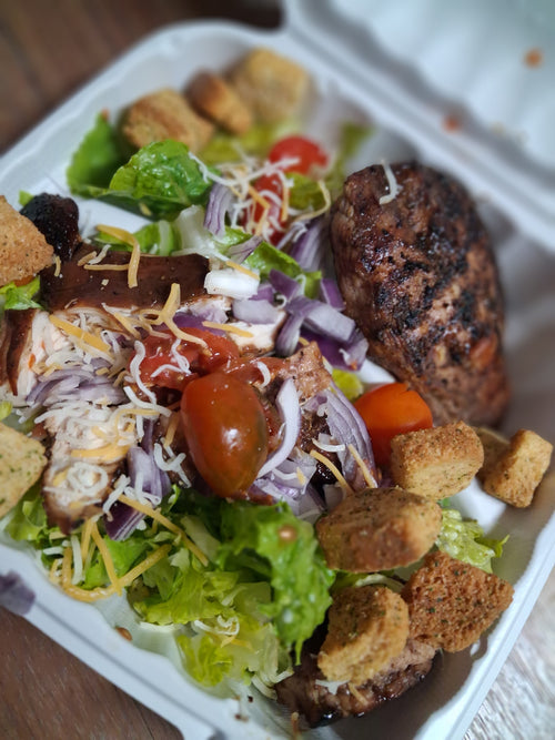 A close-up of a prepared salad in a white take-out container, featuring  grilled chicken brest mixed greens, red onion, tomatoes, shredded cheese, and croutons on a wooden surface. Prepared by Sun Connect Foods & Services LLC, Keansburg, New Jersey.