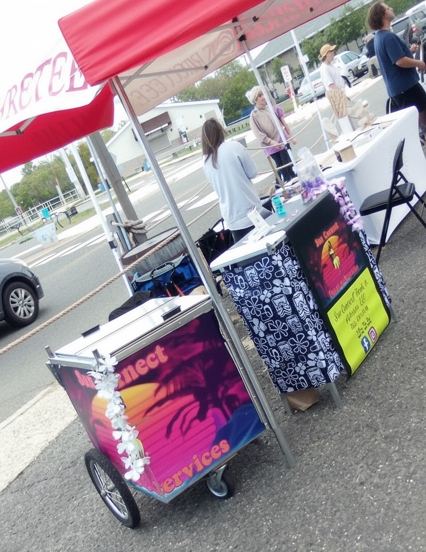 A small wheeled cart with a colorful tropical sunset graphic and the words 'Sun Connect Food & Services' on the side, photographed at an outdoor market or event next to a table with patterned fabric and signage.