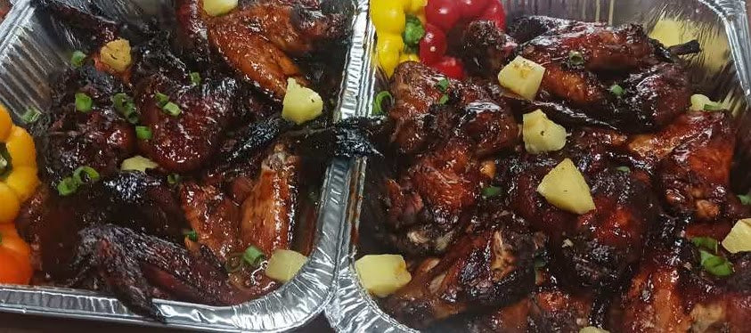 Two aluminum trays filled with glazed chicken wings and drumsticks garnished with pineapple chunks, green onions, and colorful bell pepper slices, prepared by Sun Connect Foods & Services, Keansburg, New Jersey.