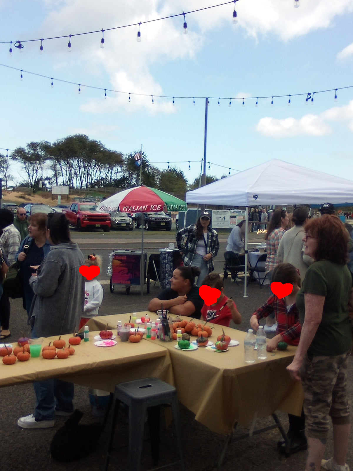 An outdoor Halloween fair scene with several people and children with red hearts drawn over their faces for privacy are gathered around tables with mini pumpkins under tents and string lights. In the background is the mobile shaved ice cart under a red, white, and green umbrella with Italian Ice written on it, by Sun Connect Foods & Services LLC, Keansburg, New Jersey